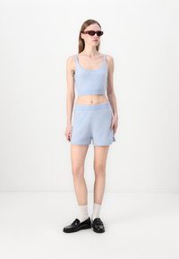Svetlomodrý pletený crop top a šortky, s rebrovanými okrajmi. Modelka má na sebe tmavé slnečné okuliare a čierne mokasíny s bielymi ponožkami.