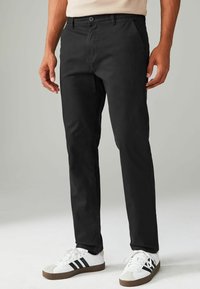 Pantalon noir en tissu lisse, avec une coupe droite, des poches inclinées, une fermeture à bouton à l'avant, associé à des baskets blanches.