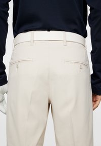 Jasno beżowe spodnie o dopasowanym kroju, z dwiema tylnymi kieszeniami, gładką teksturą i pasującym paskiem. Noszone z ciemną bluzką z długim rękawem.