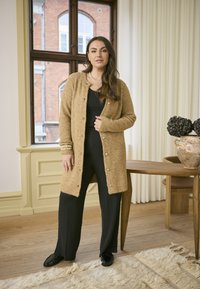 Cardigan en peluche marron clair avec des boutons, porté sur une tenue noire. Comprend des bracelets en perles et des mocassins noirs, le tout dans un intérieur moderne.