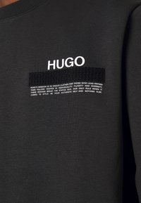 Schwarzer Stoff mit weißem "HUGO"-Text über einem schwarzen, strukturierten rechteckigen Patch und einer Missionserklärung in kleinem weißen Text darunter.