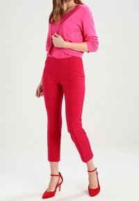 Padnoucí růžový kardigan přes růžový top, doplněný o červené kalhoty s vysokým pasem a červené lodičky na vysokém podpatku. Hladká tkanina s přizpůsobeným designem.