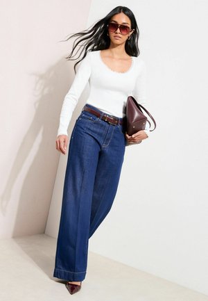 Bluză albă cu mâneci lungi și dantelă la margine, blugi albaștri închiși cu croială largă, și o geantă bordo. Model purtând ochelari de soare supradimensionați și pantofi cu toc ascuțit.