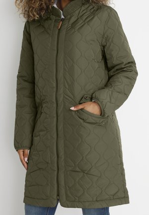 Vrouw die een olijfgroene, doorgestikte jas tot op de knie draagt met een ritssluiting aan de voorkant en zijflappzakken, met één hand in de zak, krullend haar zichtbaar.