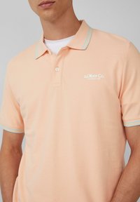 Brzoskwiniowa koszulka polo z jasnoniebieskimi paskami akcentującymi na kołnierzyku i rękawach, z wyhaftowanym logo na lewej piersi.