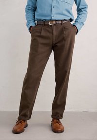 Hnědé kalhoty s dobře padnoucím střihem a předními přehyby. V kombinaci s tmavě hnědým pásem a hnědými koženými botami, na neutrálním pozadí.