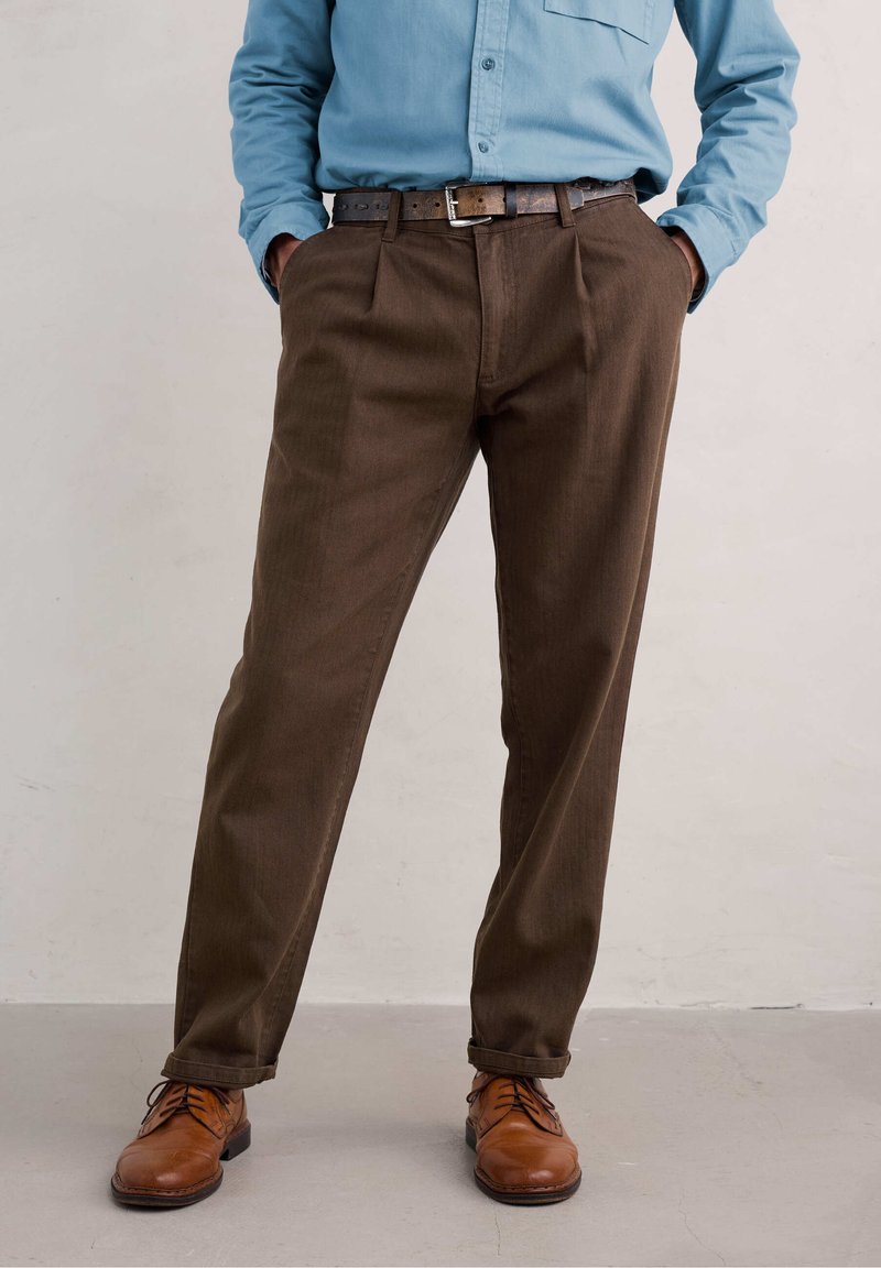 Hnědé kalhoty s dobře padnoucím střihem a předními přehyby. V kombinaci s tmavě hnědým pásem a hnědými koženými botami, na neutrálním pozadí.