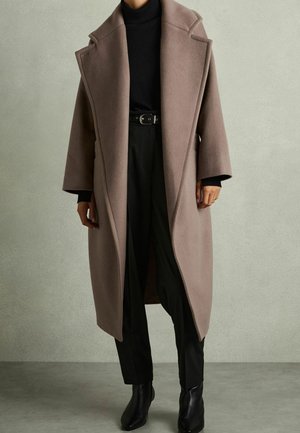 Long taupe wool coat worn over black turtleneck, black belted pants, and black ankle boots against a plain light gray background.