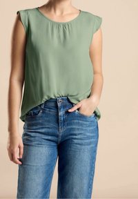 Blouse sans manches vert clair en tissu lisse avec un col froncé, associée à un jean bleu en denim taille haute avec une coupe classique.