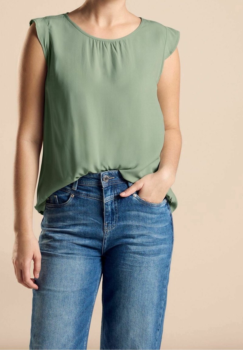 Blouse sans manches vert clair en tissu lisse avec un col froncé, associée à un jean bleu en denim taille haute avec une coupe classique.