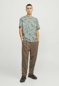Korte mouwen groene T-shirt met een wit bladdessin, gecombineerd met losse bruine broek en bruine schoenen met kwastjes. Vlakke achtergrond.