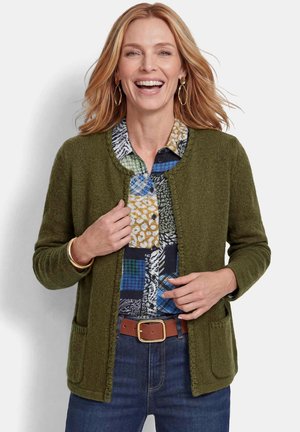 Donna sorridente che indossa un cardigan aperto verde oliva su una camicia multicolore a fantasia, jeans blu, orecchini d'oro e cinta marrone.