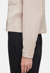 Lichtbeige jas met een gestructureerde afwerking, voorzien van een knoopsluiting bij de manchetten en een kleine metalen accent. Gecombineerd met zwarte broek.