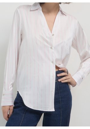 Vrouw draagt een wit overhemd met dunne roze verticale strepen en donkerblauwe spijkerbroek, hand rustend op de heup met ringen aan de vingers.