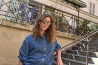 Femme portant une chemise en jean oversize et de grandes lunettes de soleil, appuyée sur une rampe près d’un escalier extérieur en pierre et d’une rambarde.