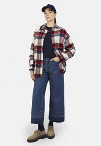 Kerman flanelli shirt in kremm med røde, sorte og gule ruter over en marineblå topp; vide blå jeans; beige sko med sorte såler; marineblå caps.