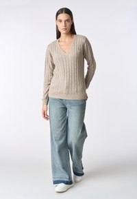 Maglione beige a trecce con scollo a V, polsini e orlo a costine, abbinato a jeans azzurri a gamba larga e sneakers bianche. Texture liscia.