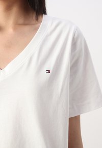 Biela bavlnená tričko s výstrihom do V, krátkymi rukávmi a malým vyšitým logom v červenej, bielej a modrej farbe na ľavej strane hrudníka.