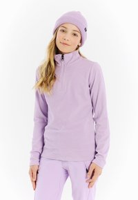 Jauna moteris vilkinti alyvų spalvos raštuotą užtrauktuką megztinį, suderintas kelnes ir megztą kepurę, stovi prieš baltą foną.