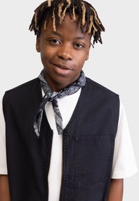 Gilet in denim nero con zip frontale e tasca con patta, indossato sopra una t-shirt bianca. Una sciarpa grigia a fantasia è annodata attorno al collo.