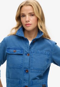 Niebieska kurtka dżinsowa z szerokim kołnierzem, trzema przednimi kieszeniami i brązowymi guzikami. Materiał ma teksturowane wykończenie, co nadaje jej swobodny, zorganizowany wygląd.