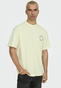 Hellgelbes Baumwoll-T-Shirt mit Rundhalsausschnitt und kurzen Ärmeln. Mit schwarzem, kreisförmigem Logo auf der linken Brust. Kombiniert mit dunklen Jeans.