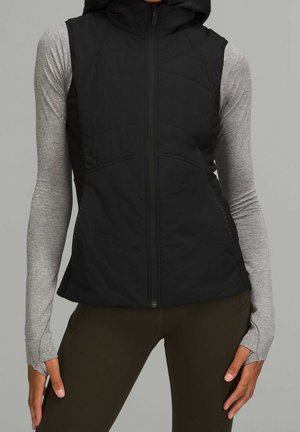 Vrouw draagt een zwart mouwloos vest met capuchon over een grijze top met lange mouwen en een donkere broek, handen ontspannen langs het lichaam op een grijze achtergrond.
