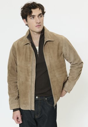 Chaqueta de ante color canela con cremallera frontal, cuello alto y dos bolsillos laterales. Se lleva sobre un top de punto marrón y vaqueros oscuros.