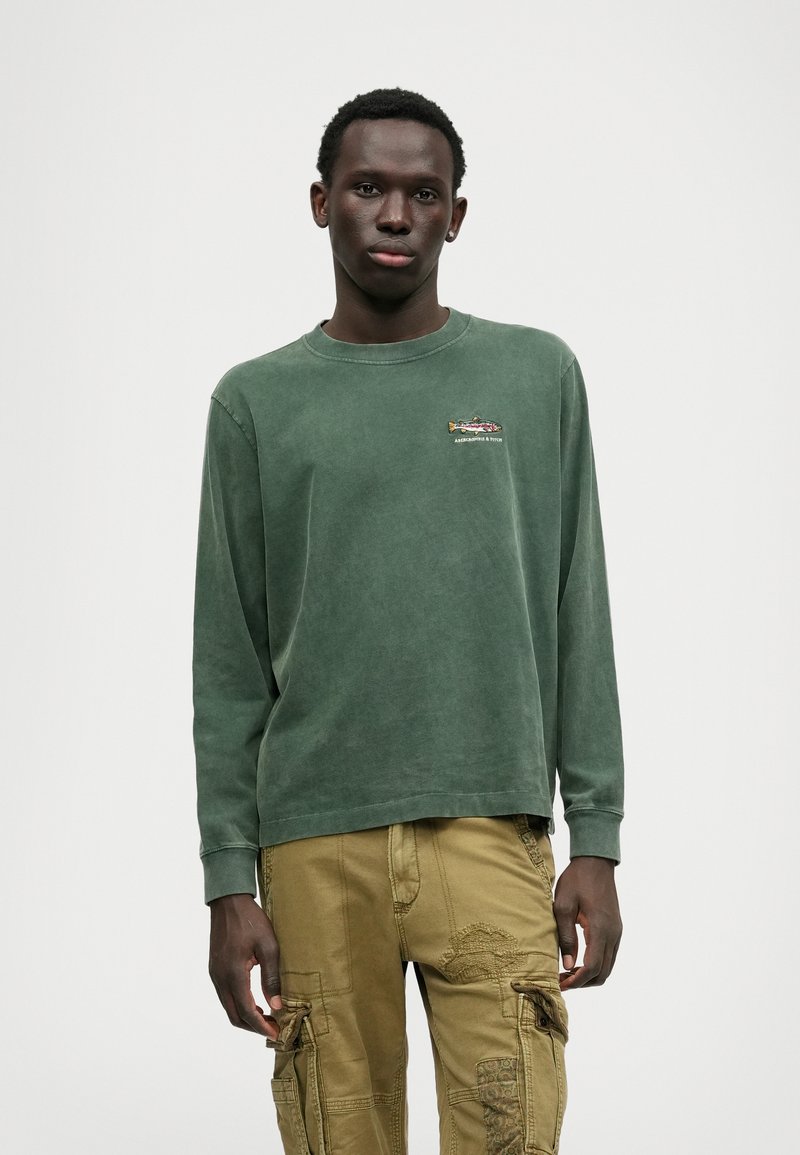 Grønn langermet skjorte laget av myk bomull, med en liten brodert design på brystet. Kombinert med khaki cargo bukser.