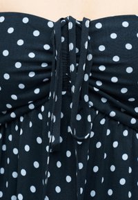 Μαύρο ύφασμα με λευκές πουά, σχεδιασμένο με ραμμένη ελαστική και δεμένες τιράντες στο κέντρο, υποδηλώνοντας το ντεκολτέ ενός φορέματος ή κορυφής.