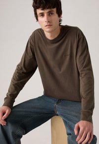 Cămașă maro cu mânecă lungă și guler rotund, având un logo subtil și asociată cu blugi din denim albastri. Textură de bumbac neted.