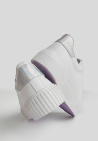 White sneakers with a glossy iridescent heel tab, ribbed sole, and textured accents. Rounded shape, fabric and synthetic materials.