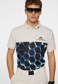 Pikétröja med en ljusbeige krage, prydd med ett svart och blått hexagonmönster. Logotyp på bröstet; i kombination med ljusa byxor.