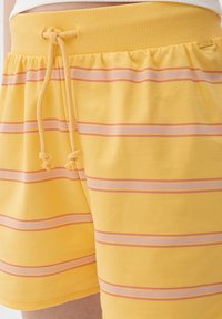 Gelbe Shorts mit horizontalen pinken Streifen, ausgestattet mit einem elastischen Bund und verstellbarem Kordelzug. Weiches Baumwollmaterial mit einer glatten Textur.