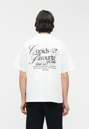 Sötét hajú személy, aki fehér pólót visel, amelyen az áll: "Cupids Favourite Victim SIMP 24/7", hátul egy angyalkép látható.