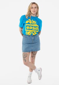 Blue graphic t-shirt with yellow "oasis" logo, paired with a denim skirt featuring vertical stripes. White sneakers and ankle socks.