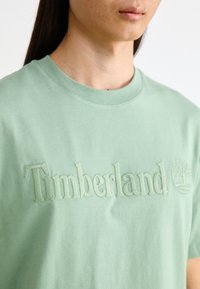 Ανοιχτό πράσινο βαμβακερό μπλουζάκι με στρογγυλή λαιμόκοψη, διακοσμημένο με το κεντημένο λογότυπο "Timberland" και το έμβλημα του δέντρου στο μπροστινό μέρος. Λείο υλικό.