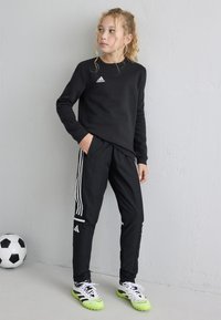 Ragazza in abbigliamento sportivo Adidas nero e scarpini neon sta vicino a un muro con un pallone da calcio sul pavimento accanto a lei.