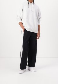 Sudadera de forro polar gris con cordón y bolsillo frontal, combinada con pantalones de pierna ancha negros con rayas laterales blancas, rematados con zapatillas blancas.