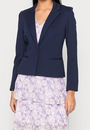 Kvinde iført en marineblå tætsiddende blazer over en lyse lilla blomsterprintet kjole med lag, stående med armene afslappet langs siden.