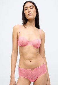 Rožinės nėriniuotos apatinio trikotažo komplektas, kurį sudaro banguota liemenėlė su lankeliais ir priekinės dalies vidurio aukščio kelnaitės, abi su geometriniais raštais ir subtiliomis detalėmis.