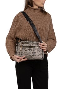 Borsa a tracolla beige con stampa del logo e dettagli neri, chiusura con zip e tracolla regolabile, tenuta da una persona che indossa un maglione di colore marrone a maglia.