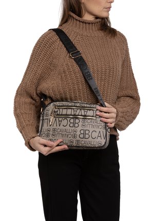 Borsa a tracolla beige con stampa del logo e dettagli neri, chiusura con zip e tracolla regolabile, tenuta da una persona che indossa un maglione di colore marrone a maglia.