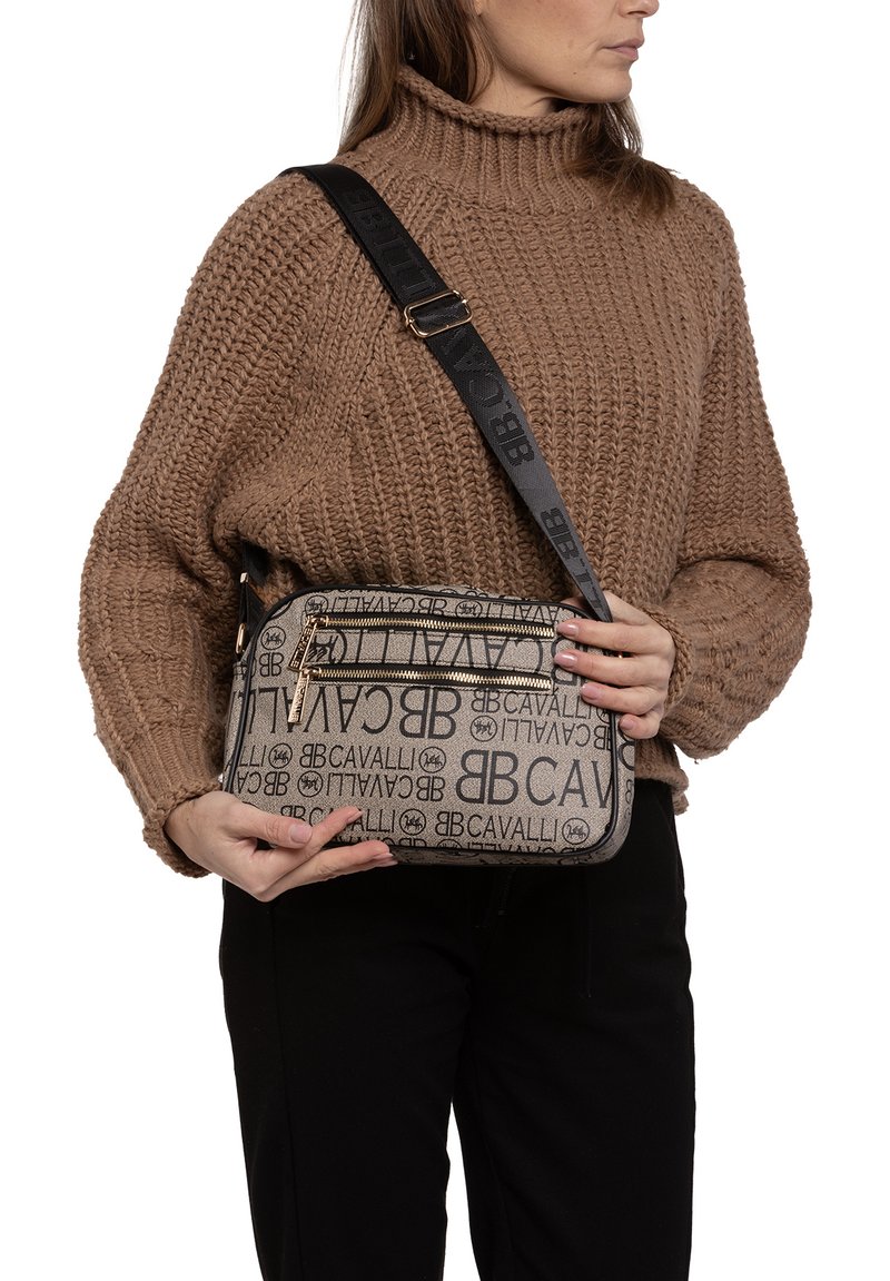 Borsa a tracolla beige con stampa del logo e dettagli neri, chiusura con zip e tracolla regolabile, tenuta da una persona che indossa un maglione di colore marrone a maglia.