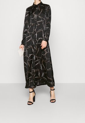 Robe noire à manches longues en tissu satiné avec un motif géométrique en blanc et beige. Associée à des talons aiguilles noirs à lanières et à une pochette à perles.