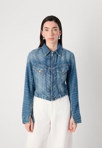 Noor naine, kellel on pikad tumedad juuksed, kannab lühendatud denimpusa ja valgeid pükse, seistes helesinise tausta vastu.