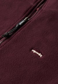 Bordowa polarowa kurtka z wysokim kołnierzem, przednim zamkiem błyskawicznym i czarnymi akcesoriami; wyposażona w małe haftowane logo przy dolnym brzegu.