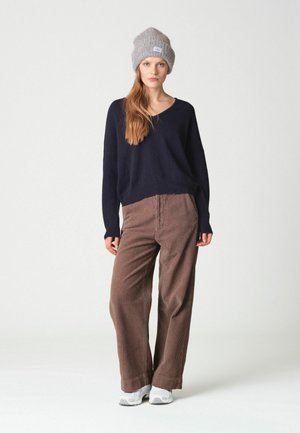 Femme portant un bonnet en tricot gris, un haut à manches longues noir, un pantalon large en velours côtelé marron et des baskets blanches, debout devant un fond uni.