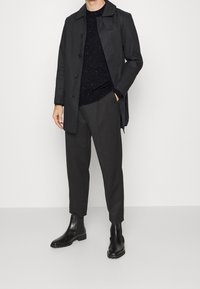 Schwarzer Außensakko mit großen Knöpfen, kombiniert mit einem strukturierten schwarzen Pullover und maßgeschneiderten grauen Hosen. Schwarze Chelsea-Stiefel vervollständigen das Outfit.