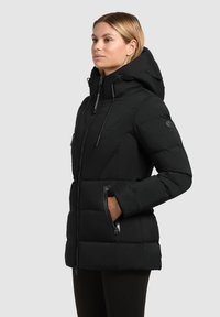 Žena v černé zateplené zimní bundě s vysokým límcem, kapucí a zipovými kapsami, stojící před jednolitým šedým pozadím.